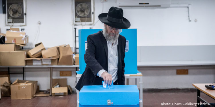 Elderly man in a dark suit and hat places a ballot into a blue voting box amidst cardboard boxes in a processing room.