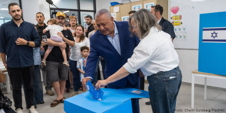Man in a blue suit dropping a ballot into a blue ballot box while a woman assists, surrounded by a diverse group of people in an indoor setting with an Israeli flag in the background.