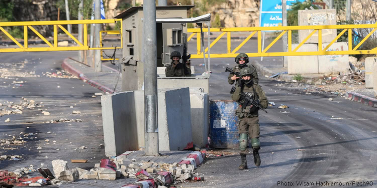 Israeli Defense Forces soldiers in tactical gear standing guard at a checkpoint with barriers and rubble in the foreground, demonstrating security operations in a conflict area.