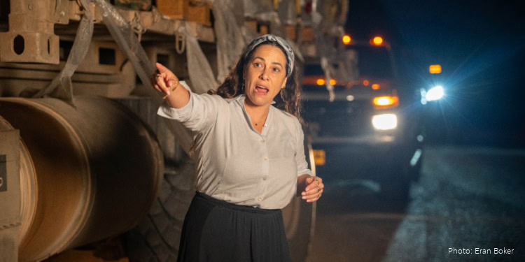 A woman in a white blouse and black skirt speaks passionately at night on a road, with trucks and cars in the background, illuminated by vehicle headlights.