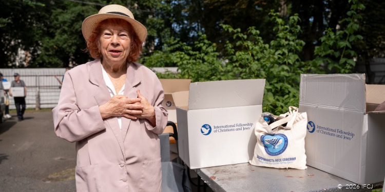 Elderly woman receiving food aid from IFCJ during a relief effort in Ukraine, with boxes and supplies on a table.