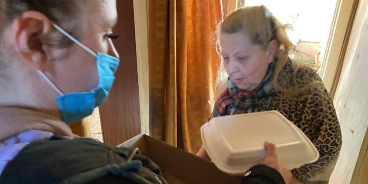 A woman in a mask delivering an elderly Jewish woman a to go box of food.
