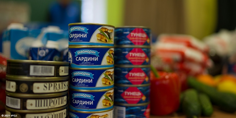 Cans of food with Cyrillic letters are stacked in front of vegetables.