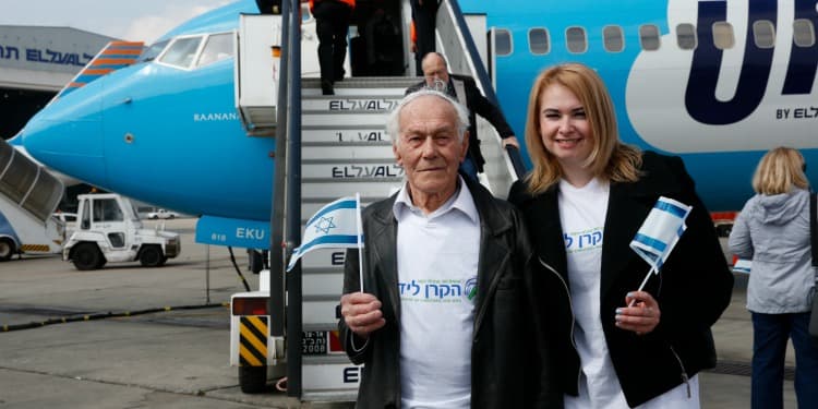Elderly man and middle-aged woman standing in front of a plane wearing IFCJ branded shirts while holding Israeli flags.