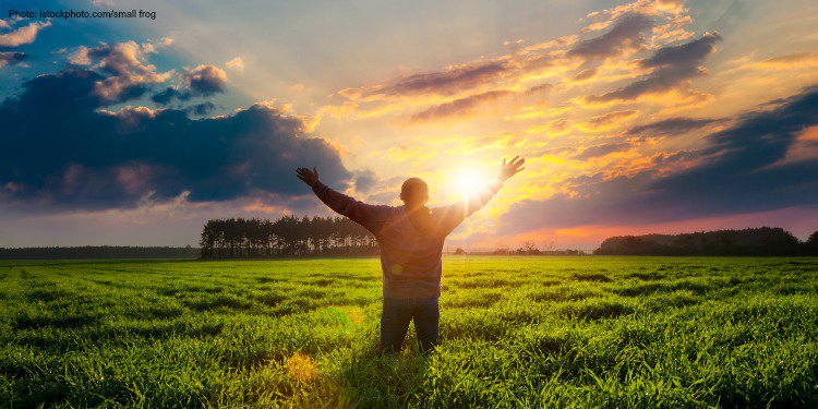 A man standing in a grassy field looking into the sunrise.