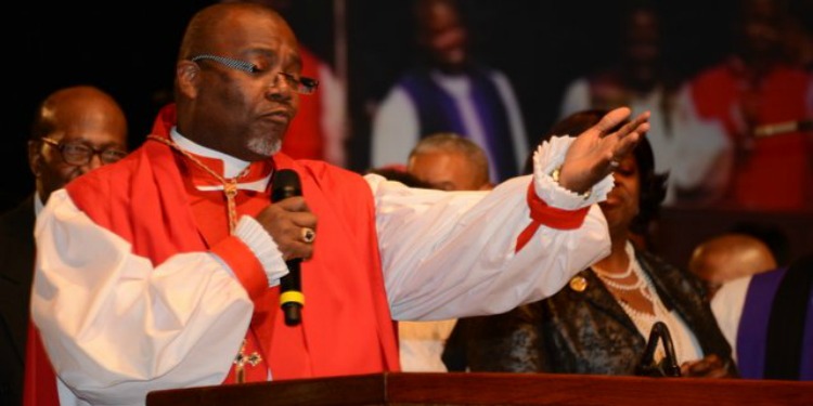 A pastor in a red and white outfit holding a microphone as he prays over the congregation.