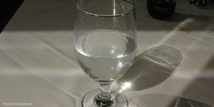 A clear glass of water placed on a white tablecloth, casting a shadow to the right, with a dark background.