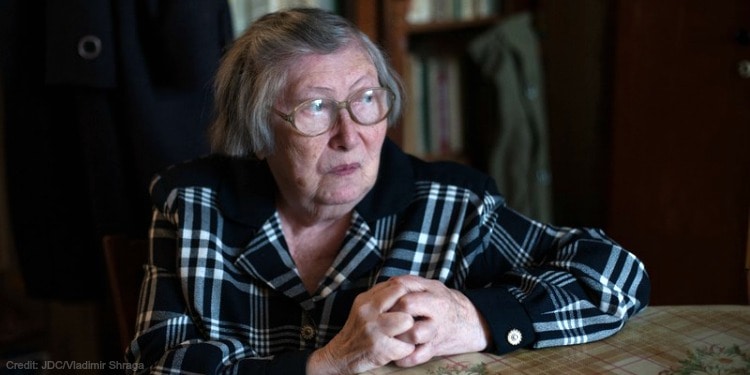 An elderly woman in glasses and a black and white patterned shirt with her hands folded on a table.