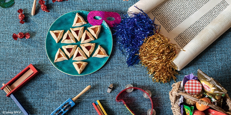 Several Purim themed things on a table including a: scroll, cookies, games, a mask, and more on a blue tablecloth.