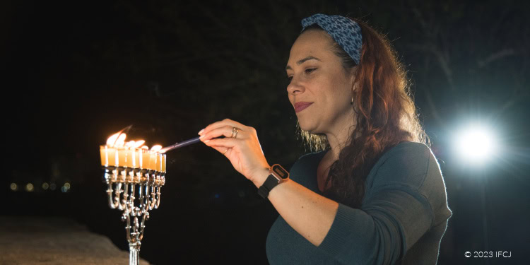 Yael Eckstein, dark green shirt, blue headscarf, smiling, happy, Hanukkah, holding candle, lighting menorah