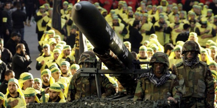 Three soldiers standing beside a rocket with a crowd of soldiers behind them dressed in yellow.