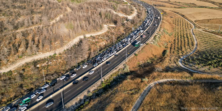 Long line of cars stuck in traffic on a rural highway surrounded by fields and hills during daytime.