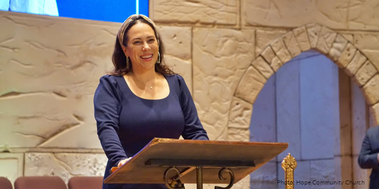 Woman speaking at a church service, smiling, with stone walls and an arch in the background.