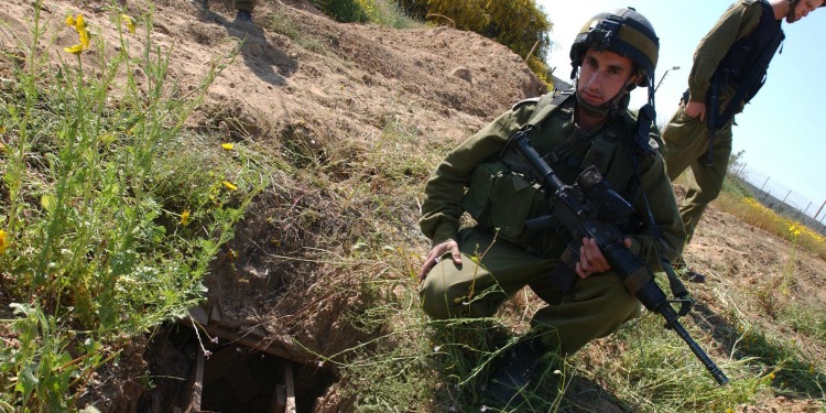 IDF soldier with a weapon squatting in the grass next to a contraption within the ground.