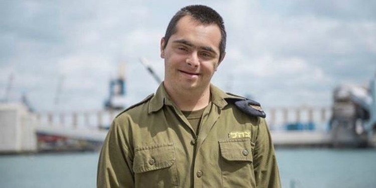 An IDF soldier that has down syndrome smiling while several machines and a body of water is behind him.