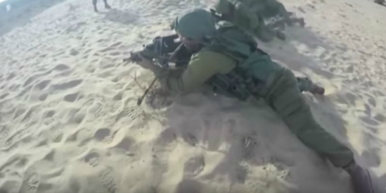 An IDF soldier laying on the sand with his weapon in a ready position.