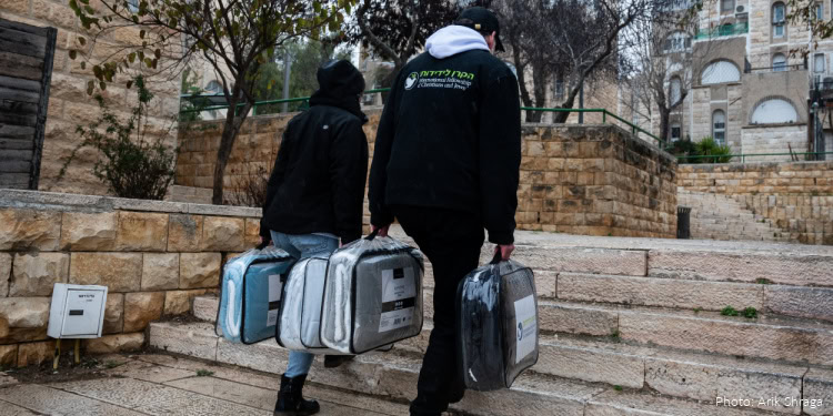 Michael Seiler (IFCJ Staff) carrying blanket in blanket case in each hand - adult man, beard, IFCJ logo black jacket, hat, glasses - young adult woman wearing black jacket - also carrying 2 blankets in blanket cases - outside, walking up steps, seen from rear