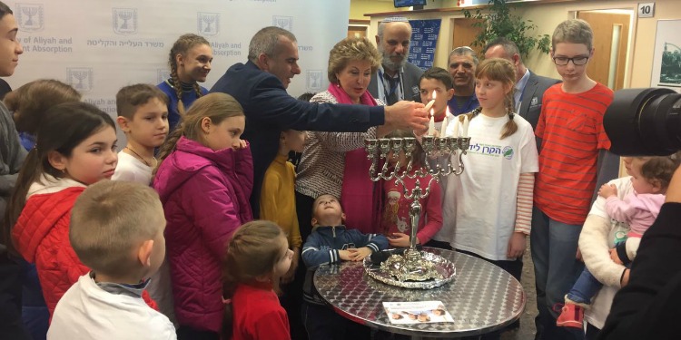 A man lighting candles on a menorah with a group of people around him, including six children who are wearing IFCJ branded shirts.