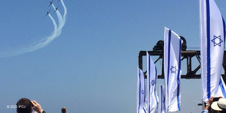 Four Israeli Air Force planes jetting off into the sky while Israeli flags are waving and people taking pictures are on the ground.