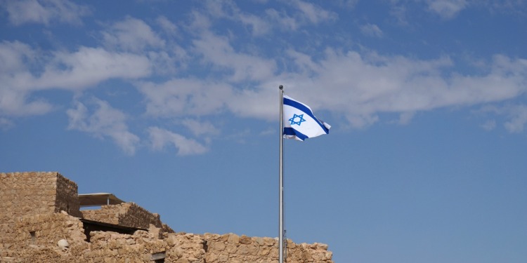 The Israeli flag on a pole waving in the sky with an old brick building behind it.