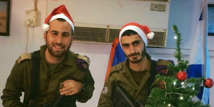 Two IDF soldiers wearing Santa hats and standing near a Christmas tree.