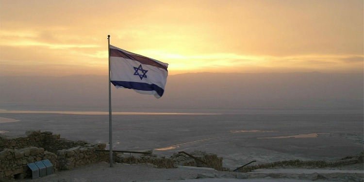 An Israeli flag waving at the top of a mountain while the sun is about to set.