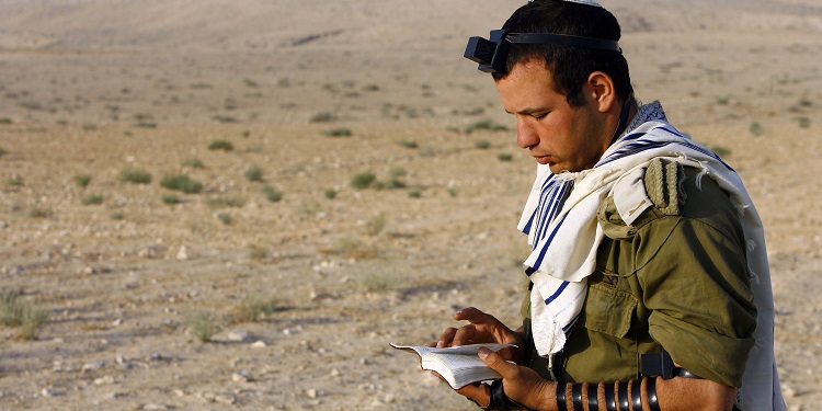 Soldier wearing tallit and tefillin prays with the Torah in the desert.