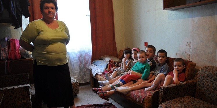 A mother standing in her living room with her six children sitting on a red couch beside her.