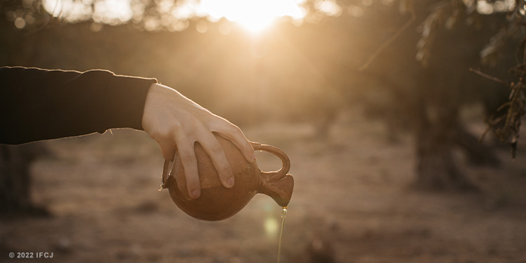 Person's arm pouring out oil from a handmade jar in a field.