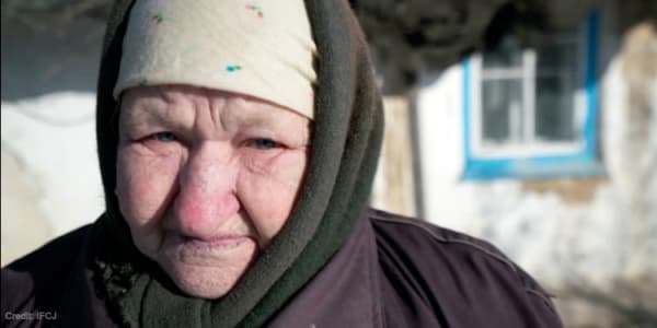 An elderly Jewish woman staring straight ahead with an old house behind her.