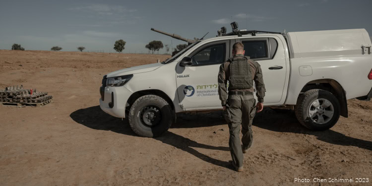 A security officer in tactical gear walks toward a white IFCJ vehicle with a mounted gun in a desert landscape, under a clear sky.