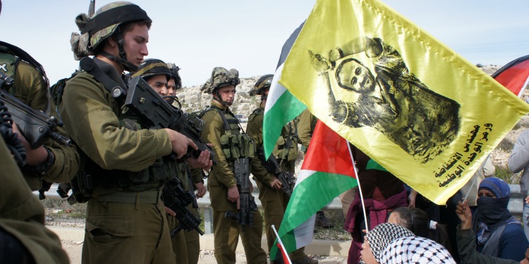 Several soldiers across from several people (mainly women and children) waving Palestinian flags.