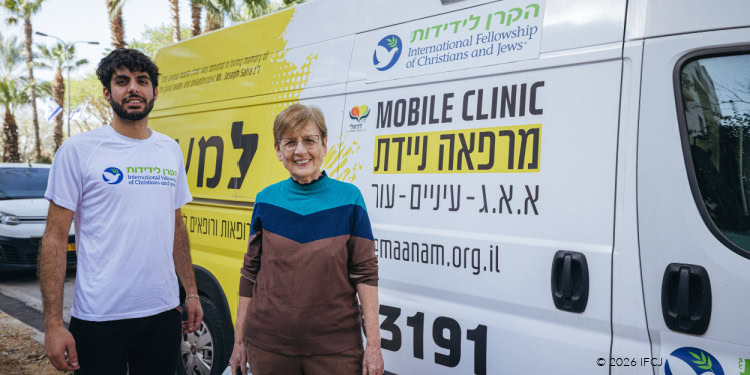 Lema’anam (For Their Sake) Medical Mobile Clinic - Visit to Pinhas and Dalia - March 11, 2026 - Israel Dr. Chaya Zehavi (ophthalmologist) and Dor Ben-Lulu (IFCJ Staff), standing in front of Lema’anam mobile medical clinic - yellow and white van - IFCJ logo - doctor - woman with glasses in brown and turquoise outfit - carrying silver metal case - man in white branded logo t-shirt