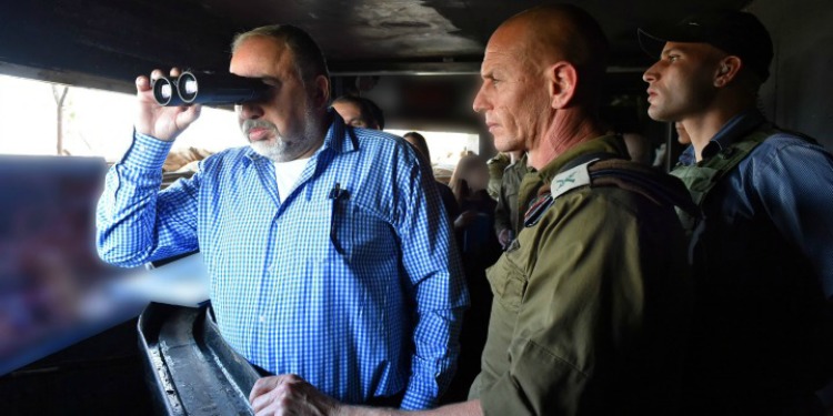 Man in a blue button down looking out from a pair of binoculars with several soldiers behind him in a bunker.