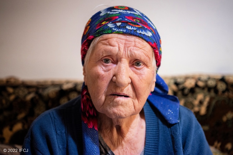 Elderly Jewish woman looking sad in blue shirt and blue scarf.