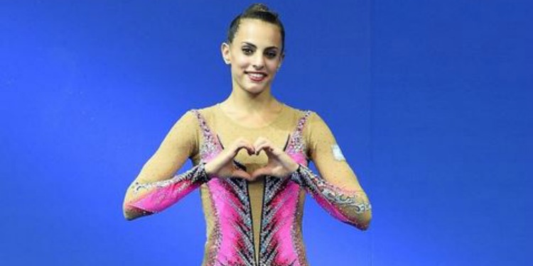 Young gymnast making a heart with her hands against a blue background.
