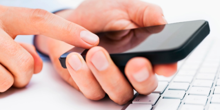 Person tapping on their phone while resting their hands on a computer keyboard.