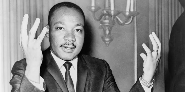 Black and white image of MLK looking into the camera while explaining something with his hands.