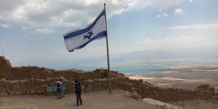 Israeli flag waving in the air against a beach background on top of a mountain.