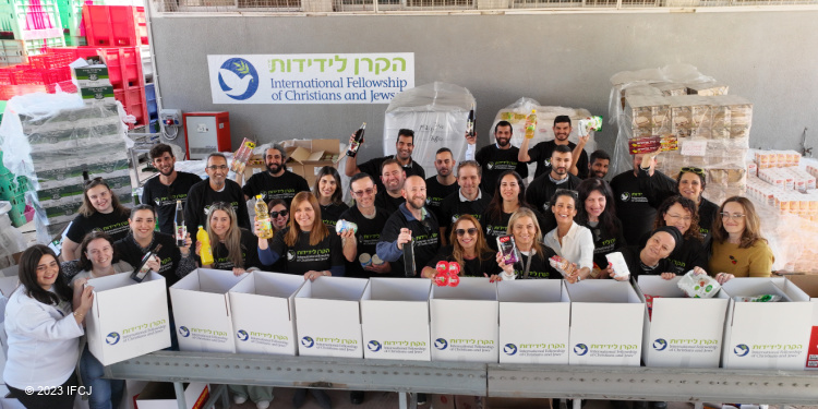 Group of IFCJ volunteers behind branded food boxes holding food in the air.