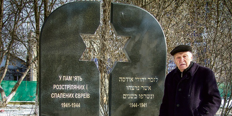 Elderly Jewish woman in a purple jacket looking into the camera while standing next to a memorial.