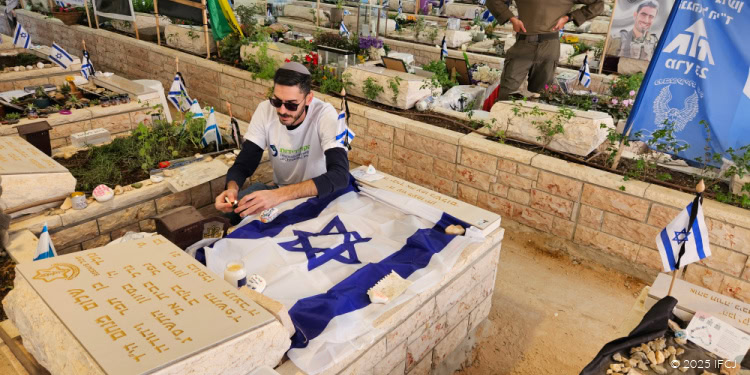 Man placing a flag on a tomb at an Israeli memorial site during a remembrance ceremony, surrounded by other graves and Israeli flags, honoring fallen soldiers.