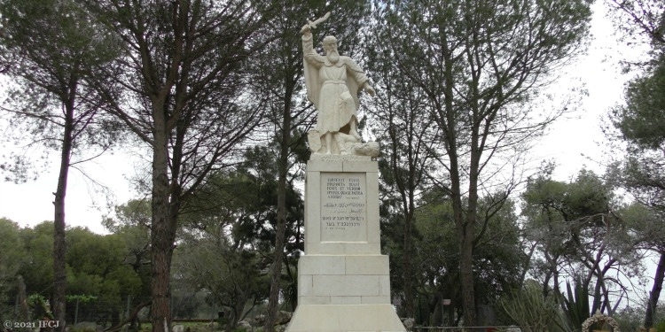 A statue of a man with a sword in his hands with trees in the background.