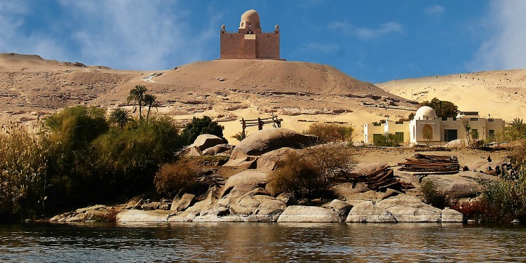 A building at the top of a sand mountain with a building below it alongside a body of water and dry shrubbery in Nile, Egypt.