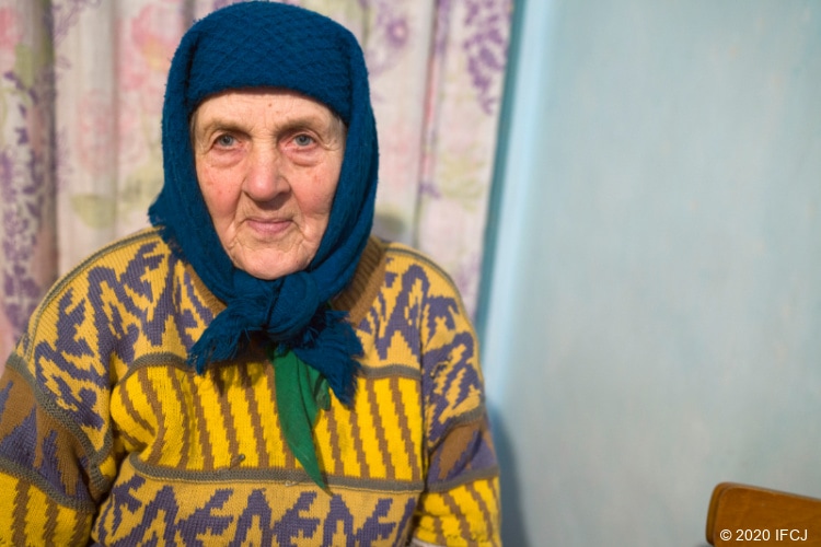 Elderly Jewish woman in yellow and blue sweater with scarf on head.