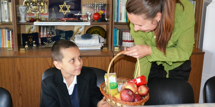 A woman giving a young child a basket filled with apples, honey, and more.