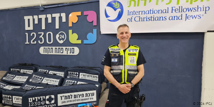 A smiling man in a reflective vest standing in front of IFCJ banners and donation boxes at a community outreach event.