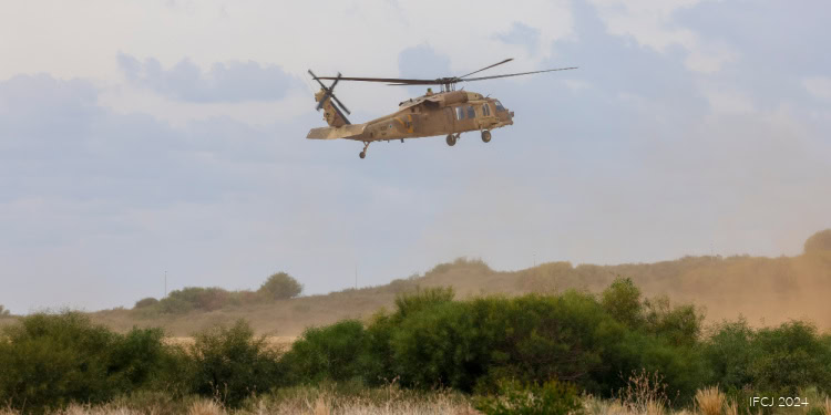 A military helicopter flying low over a green landscape with trees and bushes, under a partly cloudy sky.