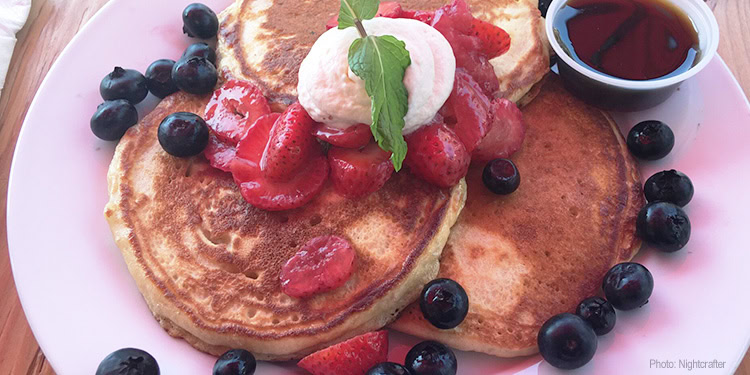 Delicious breakfast featuring fluffy pancakes topped with strawberries, blueberries, and whipped cream, served with syrup on a pink plate.