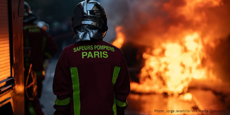 Paris firefighters in protective gear battling a large blaze with flames and smoke in the background.